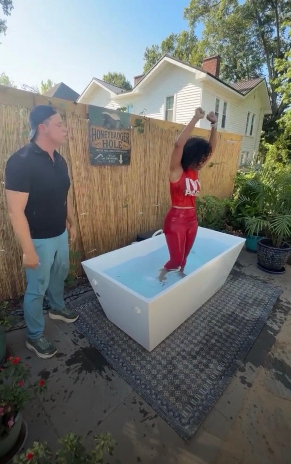 A participant raising her arms in the cold plunge tub at the Honeybadger Hole while Bill Wilder looks on